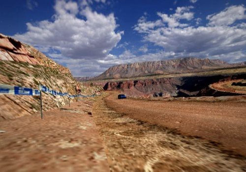 Ein blaues Rallye-Fahrzeug fährt auf einer staubigen Piste durch eine weite Canyon-Landschaft unter blauem Himmel.