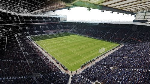 Eine weite Ansicht eines leeren Fußballstadions mit grüner Rasenfläche und Zuschauerrängen.